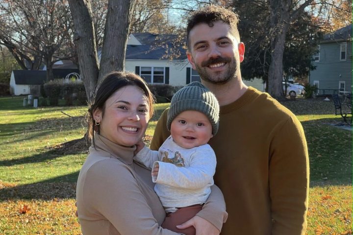 A young family of three smiles for a photo in front of their home on a sunny day. The mother wears a tan turtleneck, the father a brown sweater, and their baby a green hat and patterned shirt.