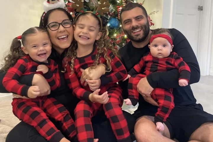 A family of five smiles in front of a Christmas tree. Two toddler girls and an infant wear matching red and black buffalo plaid pajamas. The parents wear black shirts and smile broadly.