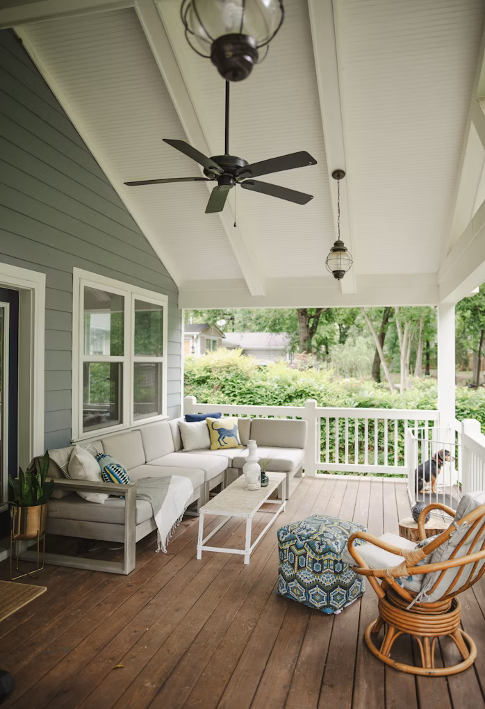 Inviting front porch with gray sectional, rattan chair, and patterned ottoman. Ceiling fan and pendant light add charm to this welcoming outdoor space.