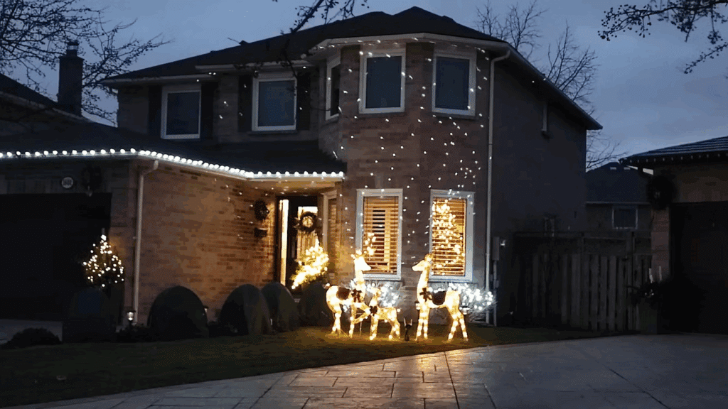 Two-story house decorated with white Christmas lights along the roofline and illuminated reindeer figures in the front yard, creating a festive holiday display.