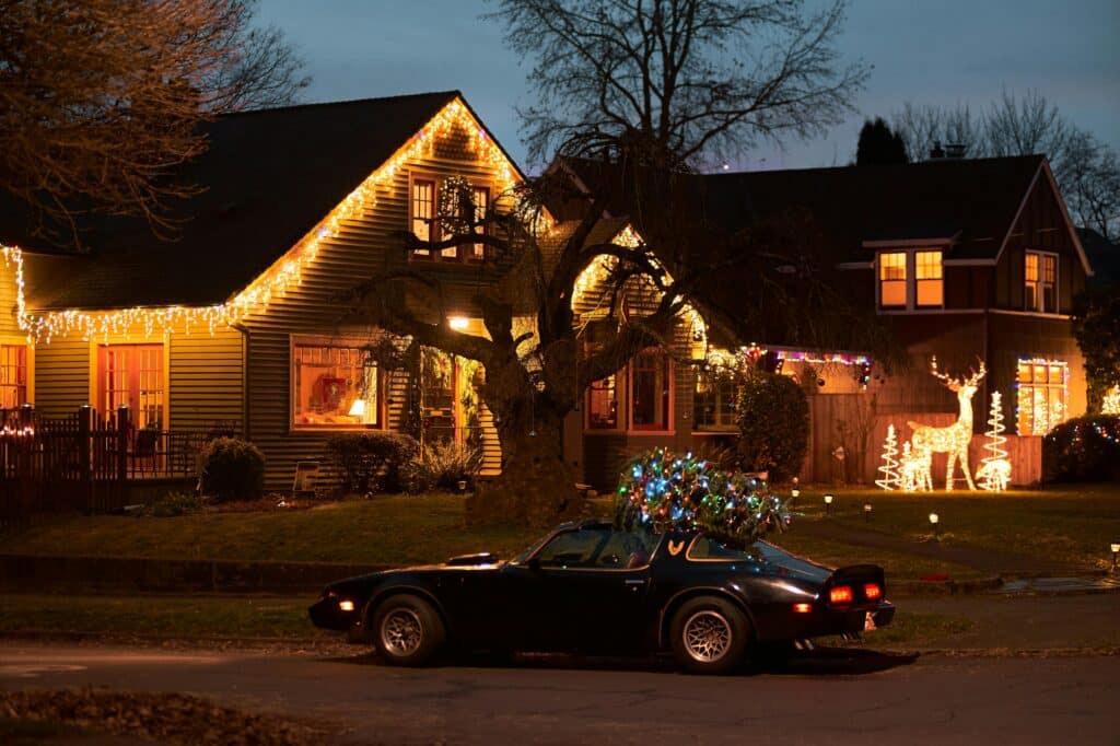 House decorated with icicle Christmas lights along the roof and yard displays including a lighted reindeer, with a car parked outside carrying a decorated Christmas tree.