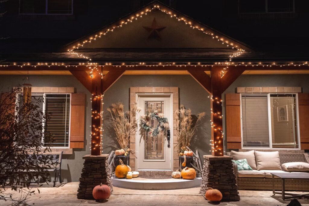 Front porch decorated with warm white string lights along the roof and columns, featuring pumpkins, dried plants, and a wreath on the door for a festive look.