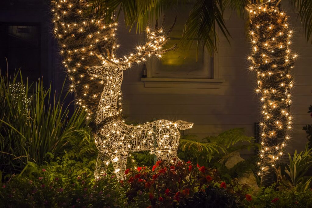 Outdoor Christmas display featuring a lighted reindeer decoration and palm trees wrapped in warm white string lights in a landscaped yard at night.